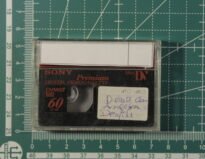 Fita MiniDV em cima de um medidor que indica que ela possui as dimensões de 5,5cm de altura e 7,5cm de largura. A fita possui uma etiqueta com o título do vídeo.