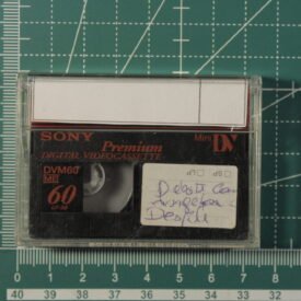 Fita MiniDV em cima de um medidor que indica que ela possui as dimensões de 5,5cm de altura e 7,5cm de largura. A fita possui uma etiqueta com o título do vídeo.